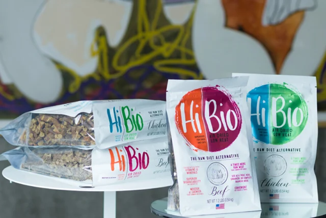 Two tables display Hi Bio freeze-dried raw diet pet food bags in beef and chicken flavors, alongside stacks of packaged pet food cubes. Abstract art is visible in the background.