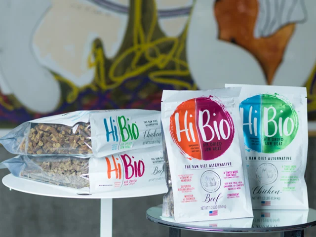 Two tables display Hi Bio freeze-dried raw diet pet food bags in beef and chicken flavors, alongside stacks of packaged pet food cubes. Abstract art is visible in the background.