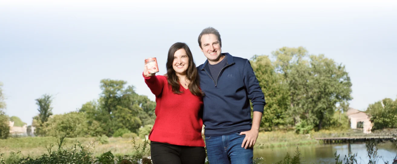Two people stand outdoors in front of trees and water. The person on the left holds up an object toward the camera, while both smile.