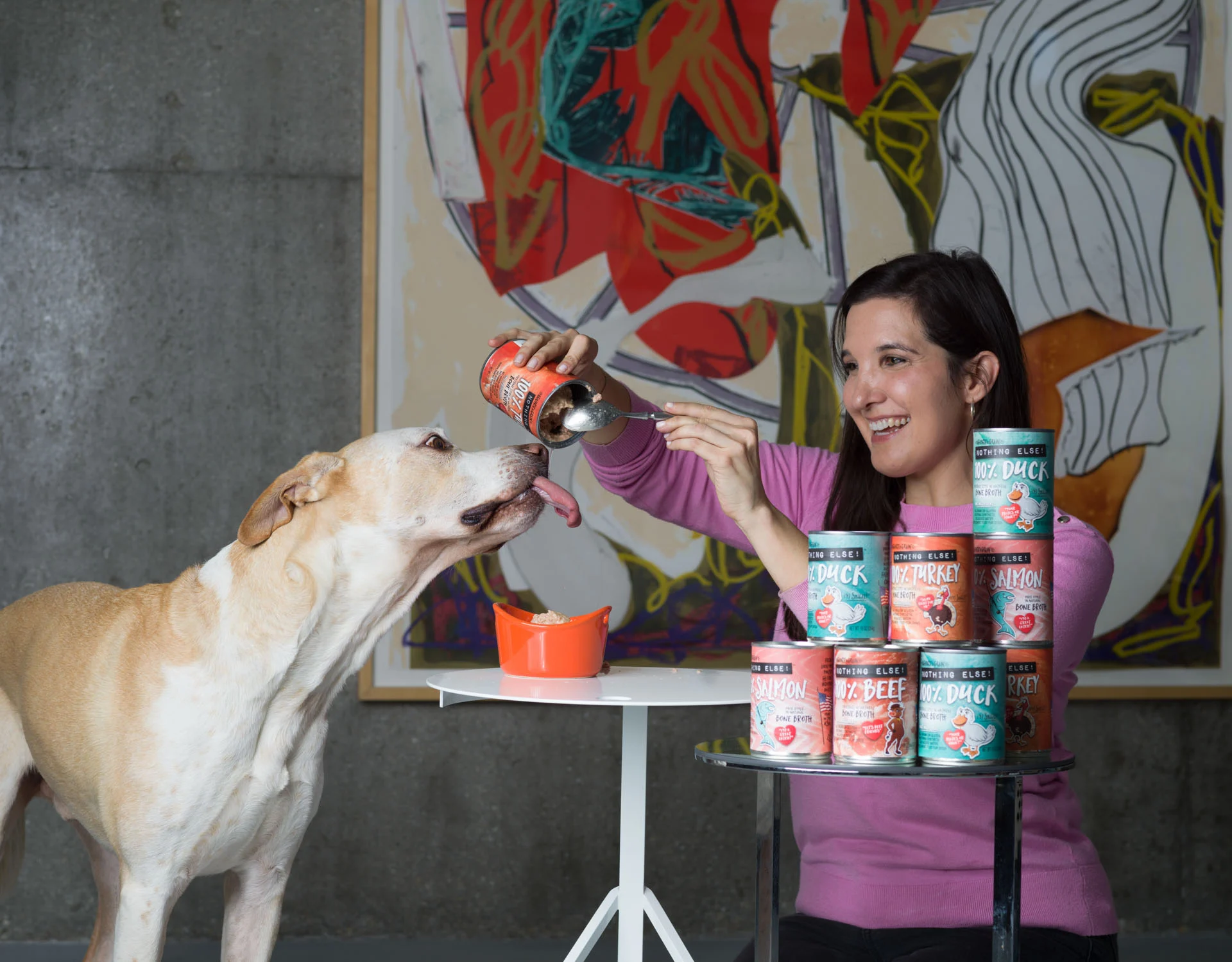 A woman feeds a large dog from a can of wet food at a table stacked with other canned pet foods. An abstract painting hangs on the wall behind them.