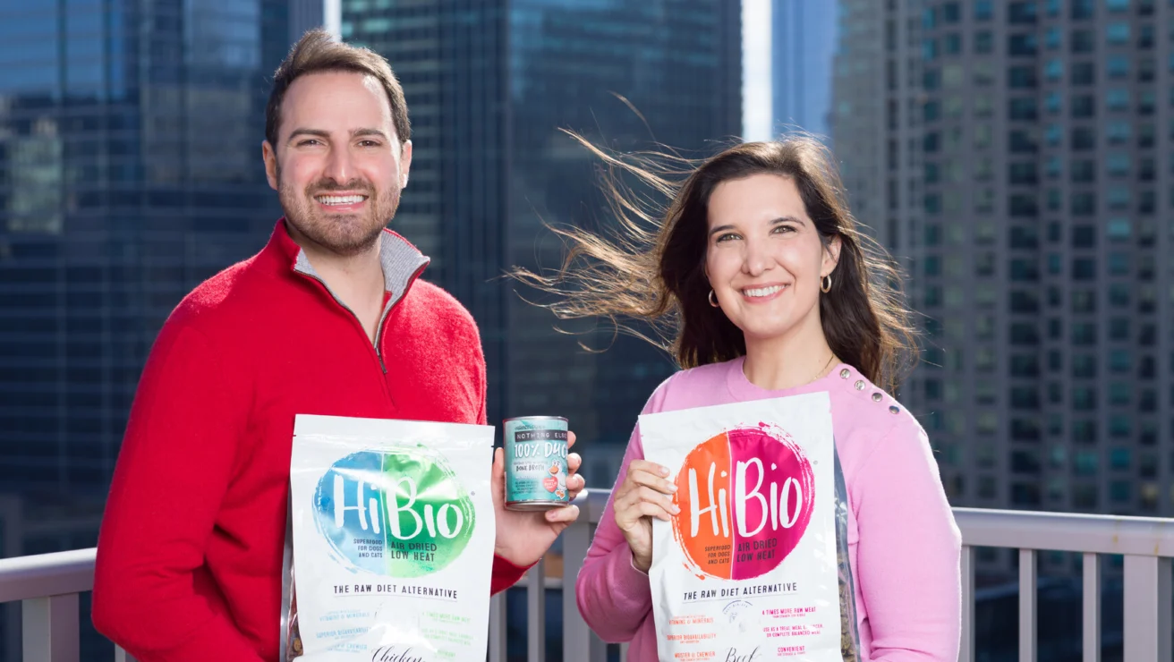 Two people stand on a city balcony holding HiBio drink products with tall glass buildings in the background.