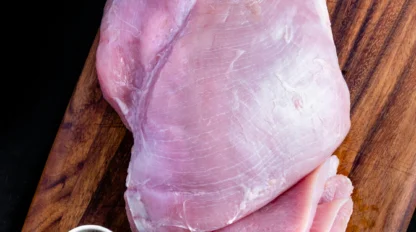 Raw turkey breast placed on a wooden cutting board with several slices cut, next to a small bowl of salt and a basil leaf.