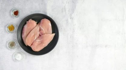 Three raw chicken breasts on a black plate, with four small bowls containing various spices, set on a light-colored surface.