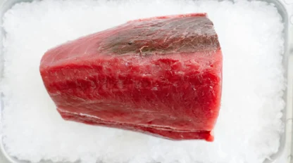 A large piece of raw tuna sits on a bed of crushed ice in a metal tray, viewed from above.