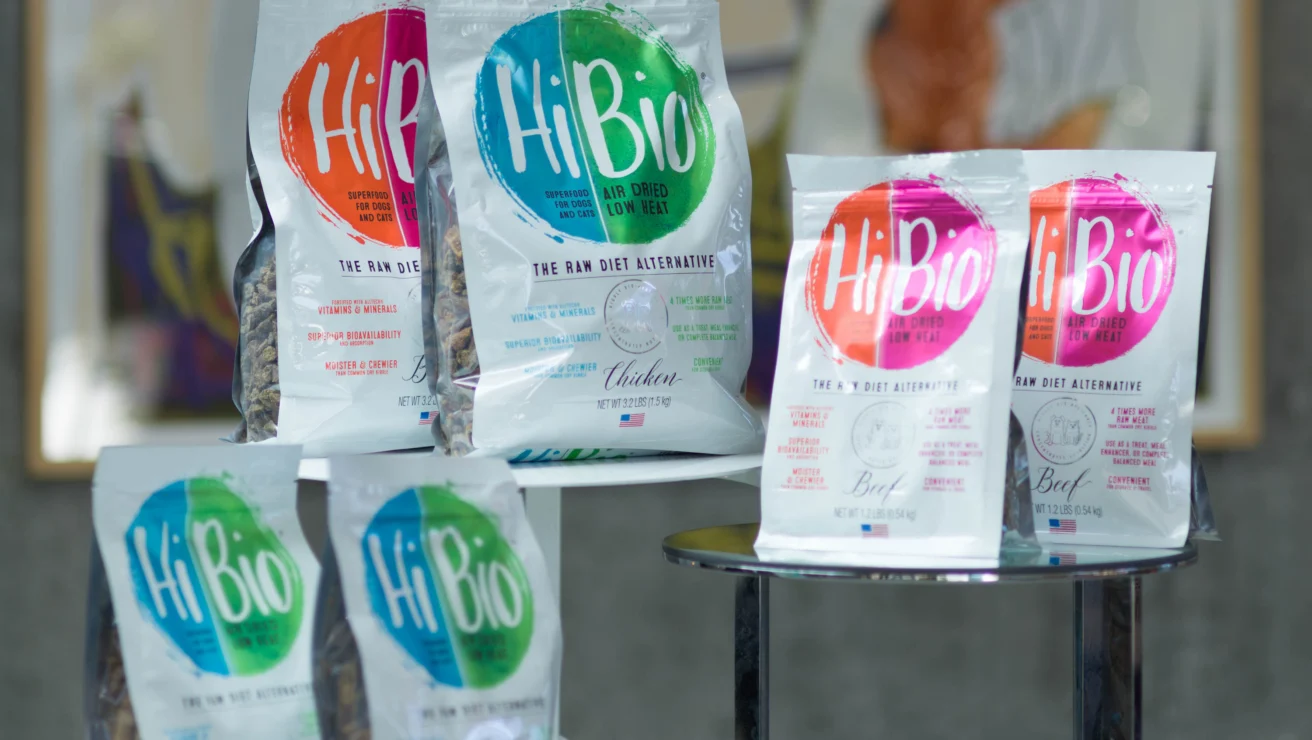 Bags of HiBio freeze-dried raw meat pet food in various flavors, including chicken and beef, are displayed on glass tables indoors.