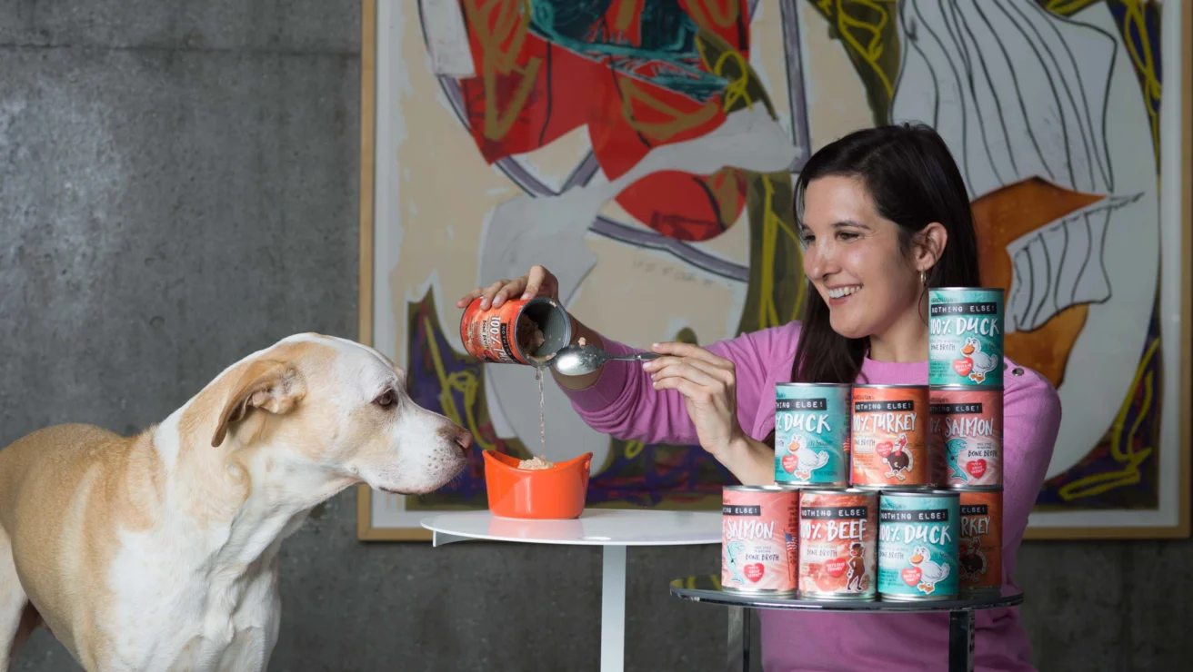 A woman pours canned dog food into a bowl for a light brown dog standing nearby. Several cans of dog food are stacked on a small table. Abstract art hangs on the wall behind them.
