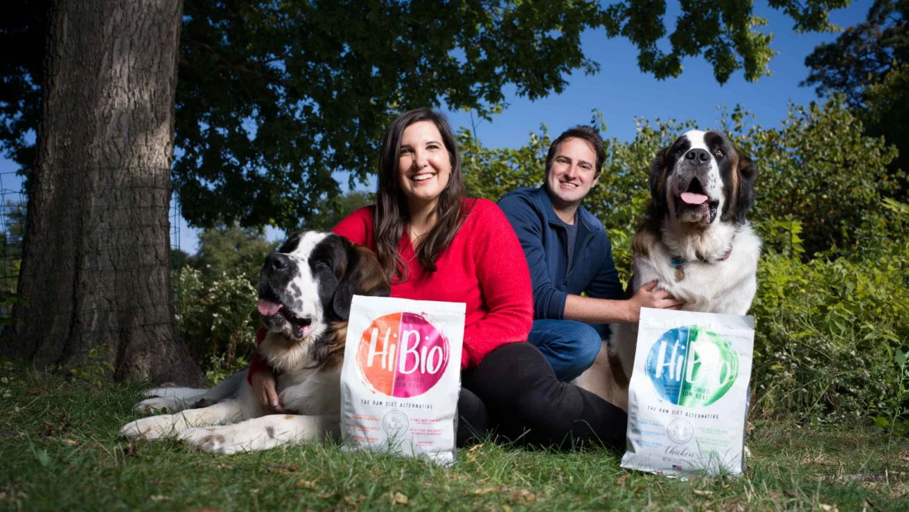 Two people sit outdoors with two large dogs, each dog resting beside a bag of HiBio dog food. Trees and greenery are visible in the background.