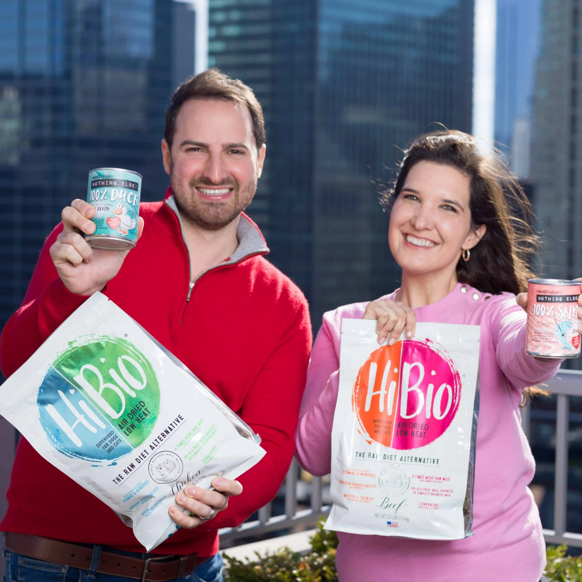 Two people stand on a rooftop holding Hi Bio pet food products and smiling. City buildings are visible in the background.